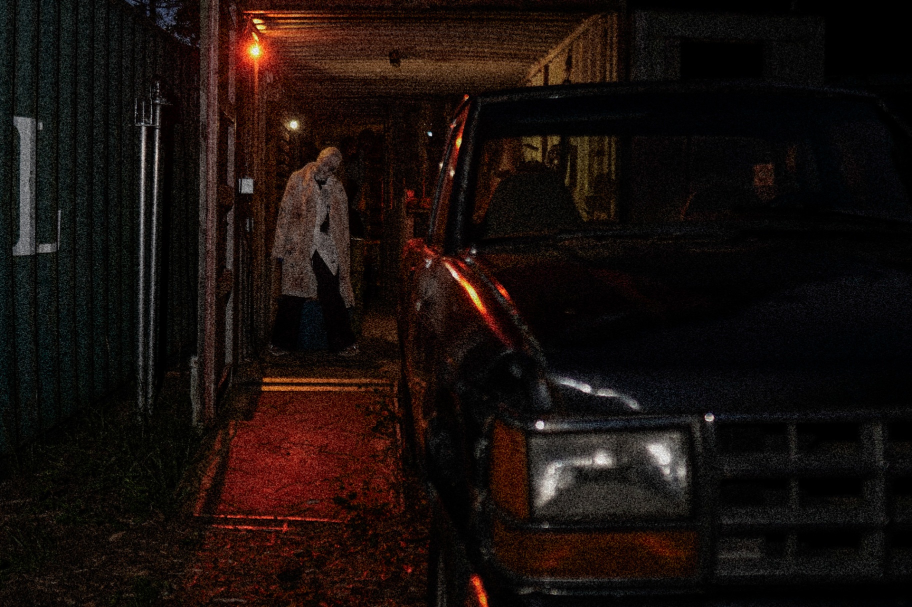 Hooded figure walking through container corridor at night beside old truck